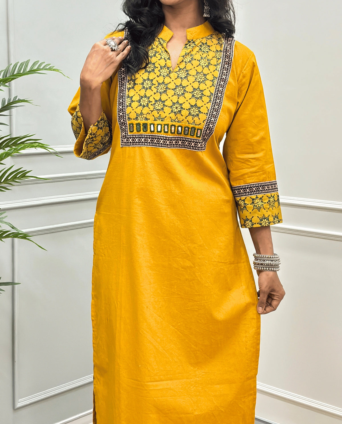 Close-up of Yellow Hand-block Ajrakh Pure cotton Long kurti with three quarter sleeves with printed cuffs,  intricate black & beige geometric embroidery, with hand mirror work and thread work on border of mirror , with Mandarian collar V-neck and sleeve cuffs, standing against a light gray paneled wall. 