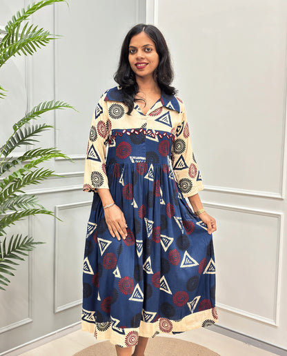 Navy-blue Women's Ajrakh Vegetable dyed plant based Cotton Long-Kurti with a Beige neck and cuffs, and a Navy blue -colored collar & dress featuring a geometric pattern of triangles and spiral in beige, black, and Red, Hand-mirror work on front, standing on a round woven rug in front of a white paneled wall with a green potted plant on the left.