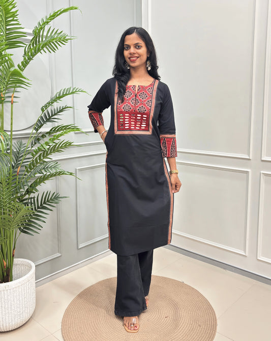 Black Women's Hand-block Ajrakh Pure Cotton Long Kurti with a red base and abstract Black & Beige  geometric embroidery on the chest and sleeve cuffs, paired with black Flared pants, standing in a decorated room with a potted plant.