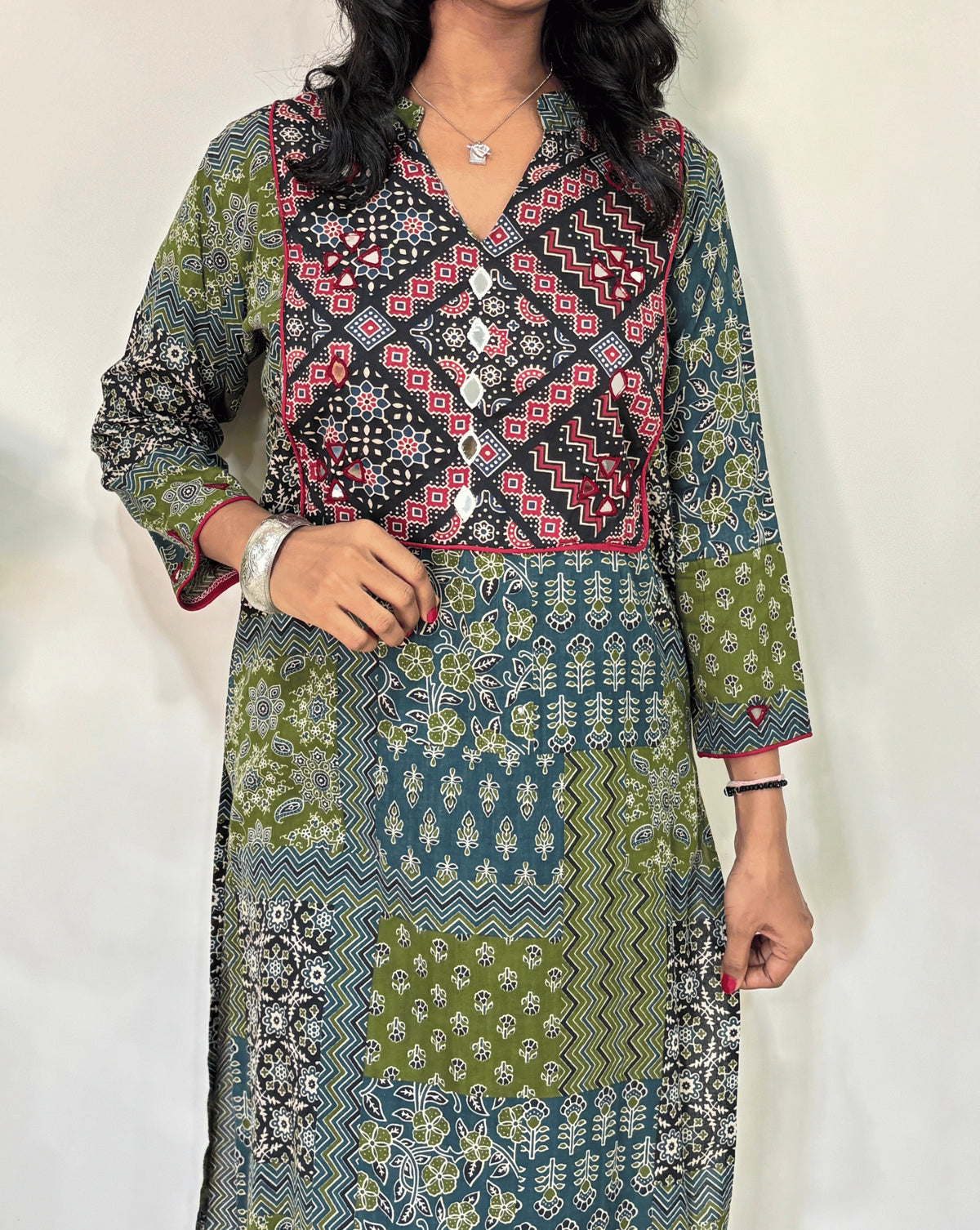 Close-up of Women's Green Ajrakh Vegetable Dyed plant based cotton long kurti Featured with full sleeves, with green, black, and red patterns, Hand-mirror work on front & Sleeves , with Mandarian V-shaped neck, paired with black flared pants, standing in a room with a white wall and a potted plant.