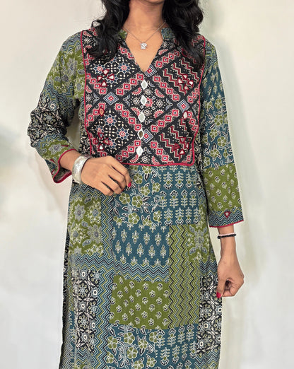 Close-up of Women's Green Ajrakh Vegetable Dyed plant based cotton long kurti Featured with full sleeves, with green, black, and red patterns, Hand-mirror work on front & Sleeves , with Mandarian V-shaped neck, paired with black flared pants, standing in a room with a white wall and a potted plant.
