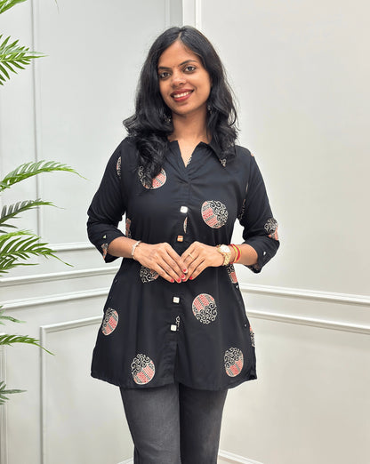 Front view of Black Women's Ajrakh Vegetable dyed Plant based A-line cotton Short kurti with pink & white floral embroidery, Hand-mirror work on front look a like buttons , Sleeves Cuffs & pockets, paired with black flared jeans, standing on a round woven rug in a modern room with a potted plant.