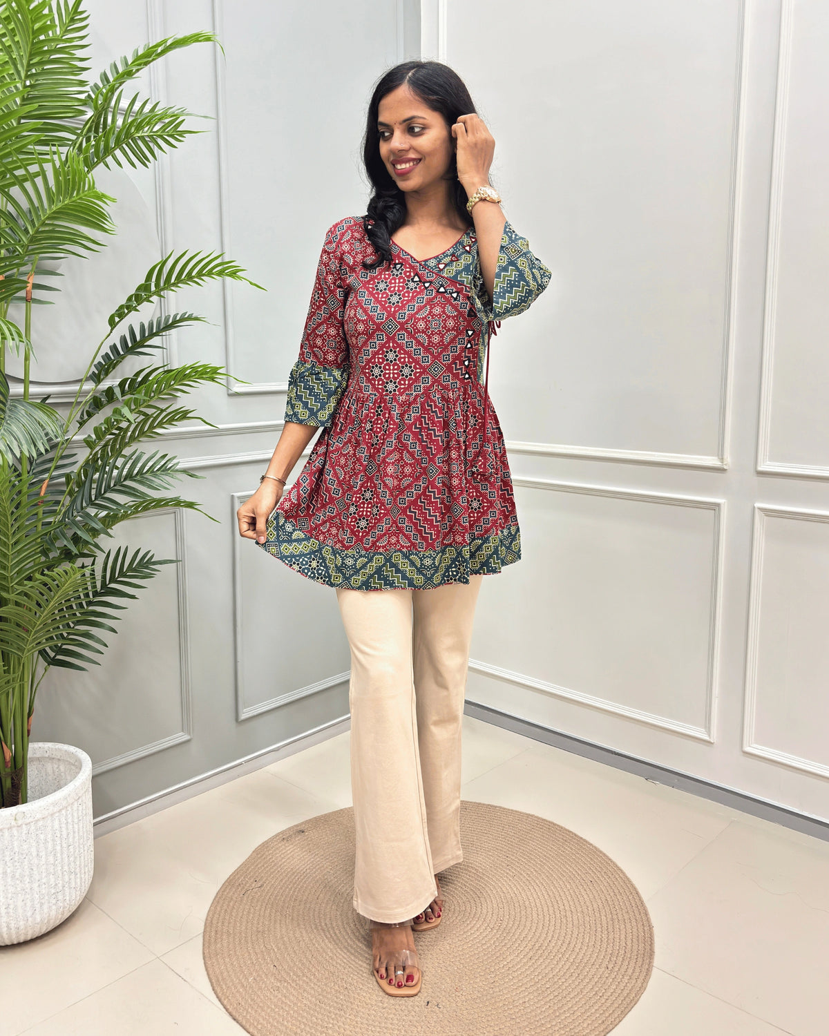 Red Women's Ajrakh Vegetable Dyed plant based cotton Short Kurti with a teal and Beige geometric pattern, paired with beige flared pants, standing on a round woven mat in front of a white paneled wall with a potted plant.