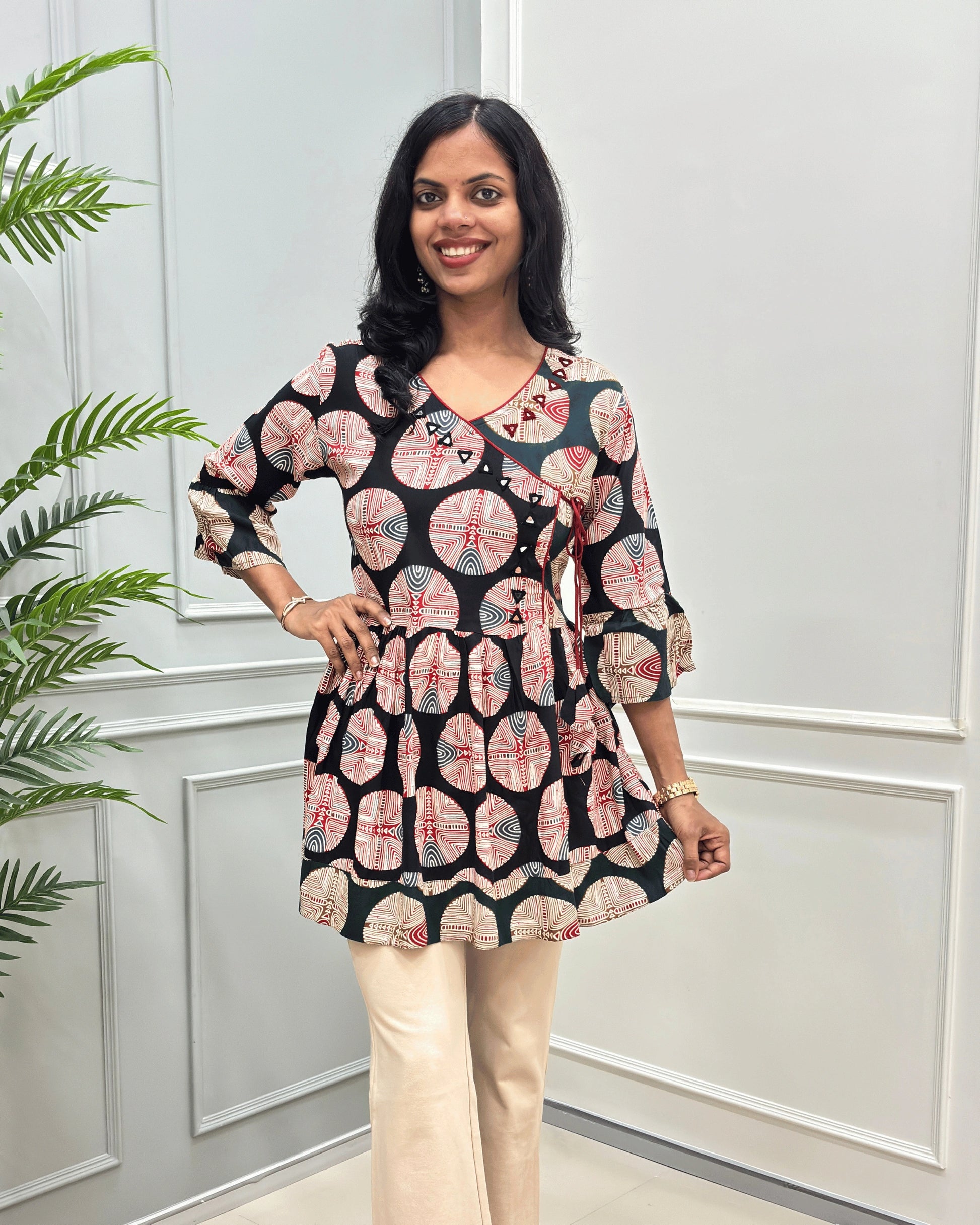 Front view of Black women's Ajrakh Plant based cotton Short-Kurti with a multicolor abstract print of circles and geometric patterns, Green color base on top bottom & sleeves cuffs in paired with beige pants, standing in front of a white paneled wall.