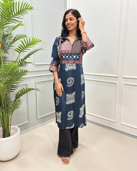 Navy-Blue women's Pure Mashru Silk Long Kurti with red , Black & white geometric patterns on the yoke and sleeves, paired with black wide-leg pants and brown sandals, standing in a room with a potted plant.