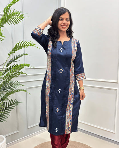 Front view of Navy Blue Ajrakh Mashru Silk Long kurti with intricate Multi-color embroidery on the front panels and cuffs,with hand mirror work,  paired with maroon pants, standing in a decorated room with a potted plant.