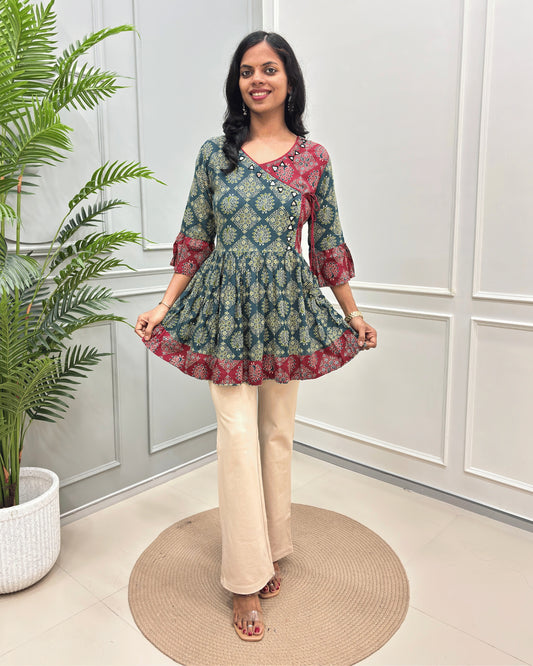 Green Women's Ajrakh Vegetable Dyed plant based cotton Short Kurti with a teal and red geometric pattern, paired with beige flared pants, standing on a round woven mat in front of a white paneled wall with a potted plant.