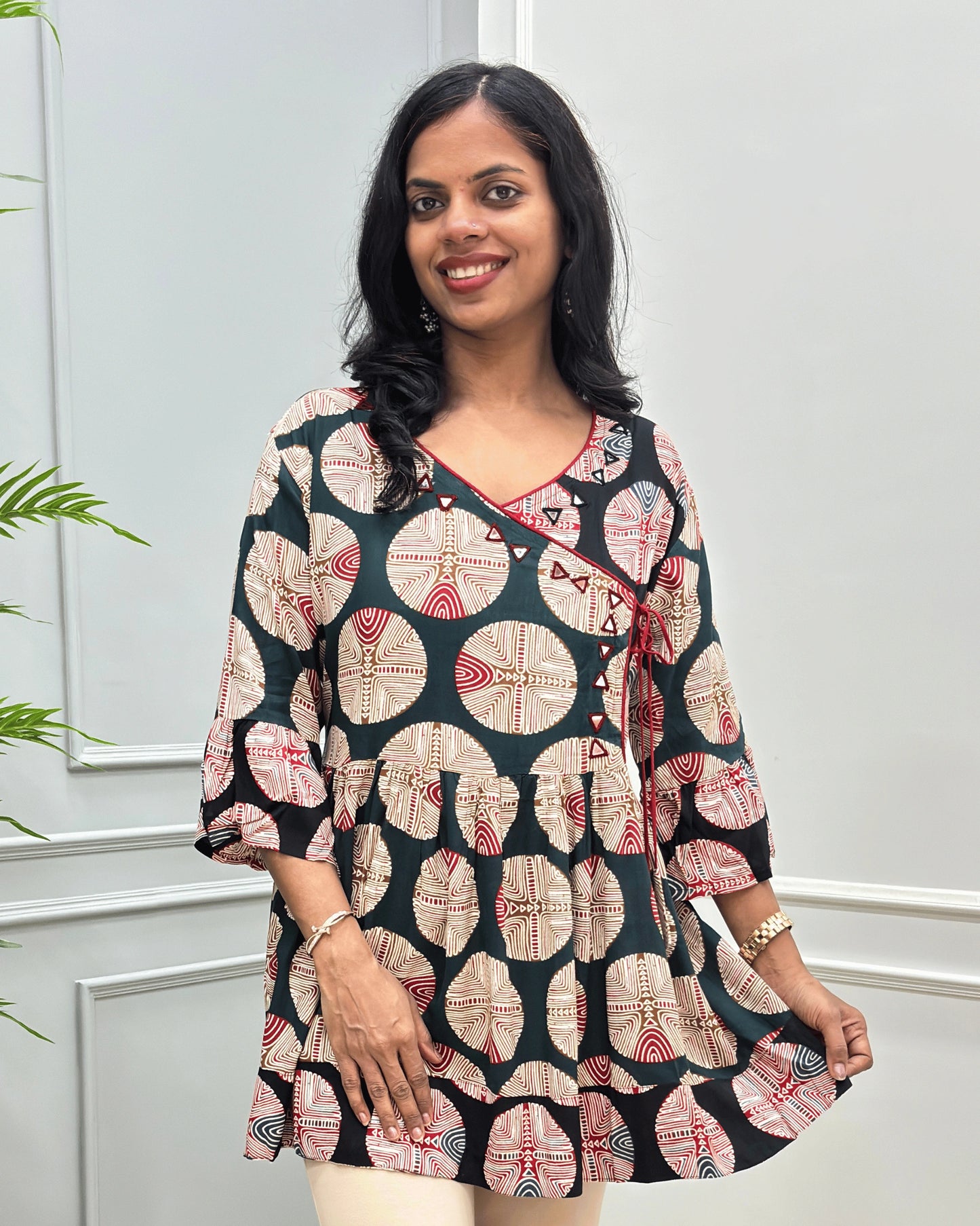 Close-up of Green women's Ajrakh plant based cotton Short kurti with a colorful geometric and circular print, Hand-Mirror work on V- neckline along with lace, paired with beige pants, standing in front of a white paneled wall with a green plant on the left.