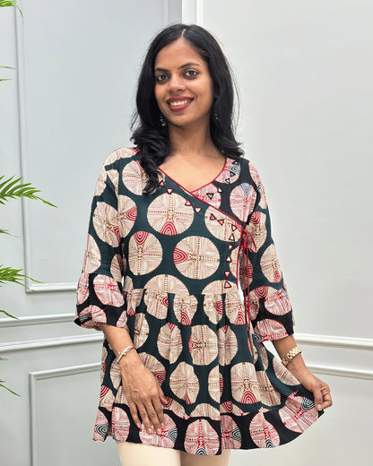 Close-up of Green women's Ajrakh plant based cotton Short kurti with a colorful geometric and circular print, Hand-Mirror work on V- neckline along with lace, paired with beige pants, standing in front of a white paneled wall with a green plant on the left.