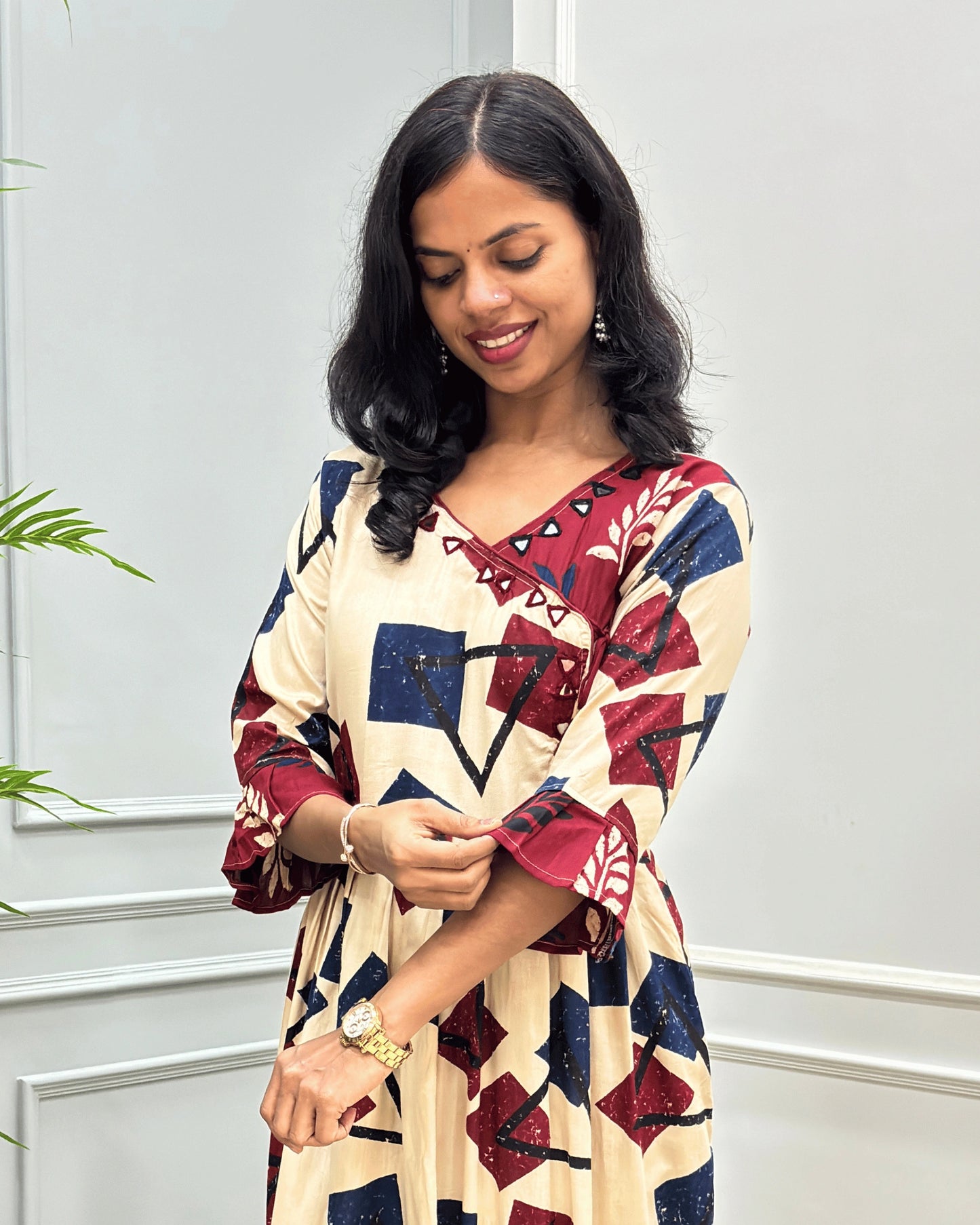 Close-up of beige women's plant based cotton long-kurti  in navy blue and red, with leaf print on calf of frill sleeves, hand-mirror work on V-shaped neck and standing against a light gray paneled wall