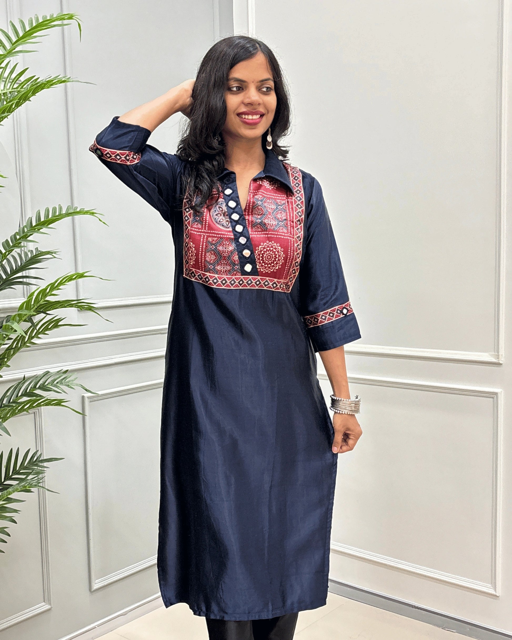 Front view of Navy-blue women's Ajrakh Mashru Silk with three-quarter Sleeves Featured with detailed multicolor embroidered yoke and patterned sleeve cuffs with Bandhani print , standing in front of a white paneled wall with a green plant on the left.