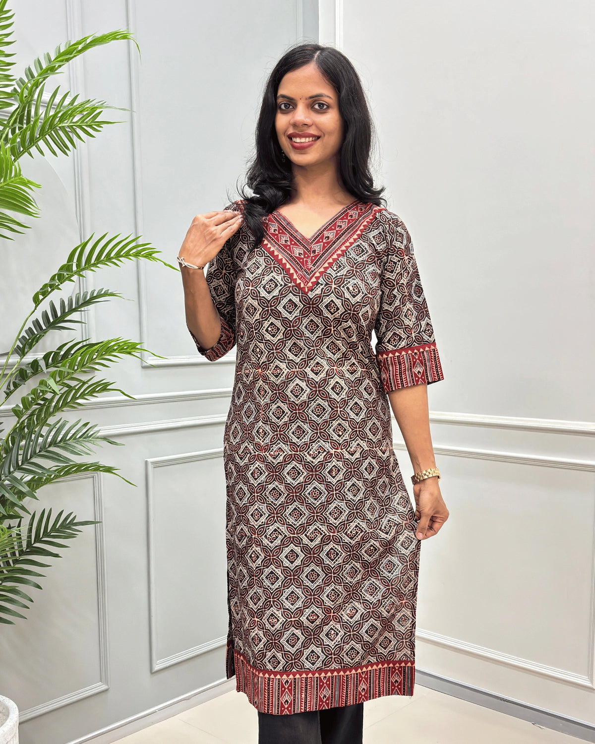 Front View of Black Women's Ajrakh Hand-block Cotton Long Kurti with red and Black ajrakh Print on V-neck ,Strip print on trim & Sleeves , standing in front of a white paneled wall with a green plant on the left.