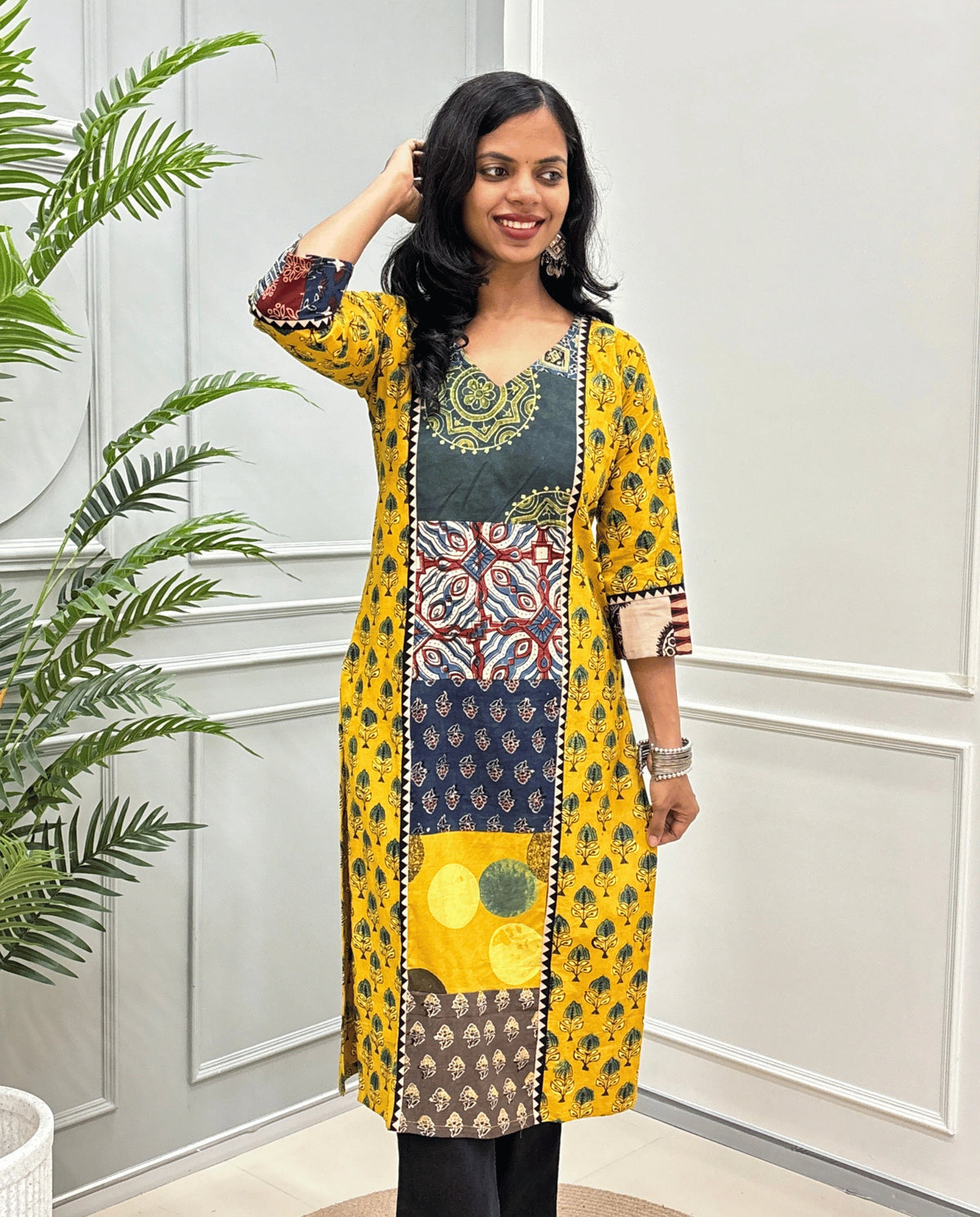 Front view of Yellow women's Hand-block Ajrakh pure cotton long kurti with three quarter full printed sleeves with a yellow base, & Abstract red, yellow, Brown and black Ajrakh Geometric print panel & cuffs, standing in front of a light gray paneled wall with a green plant on the left.