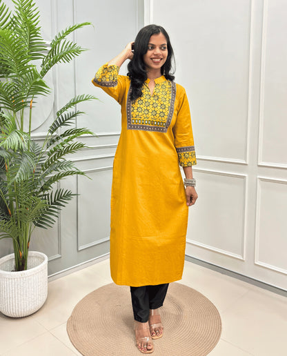 Yellow Women's Hand-block Ajrakh Pure Cotton Long kurti with intricate Green geometric print on the chest and sleeve cuffs, paired with black pants , standing on a round woven mat in front of a white paneled wall with a potted plant.