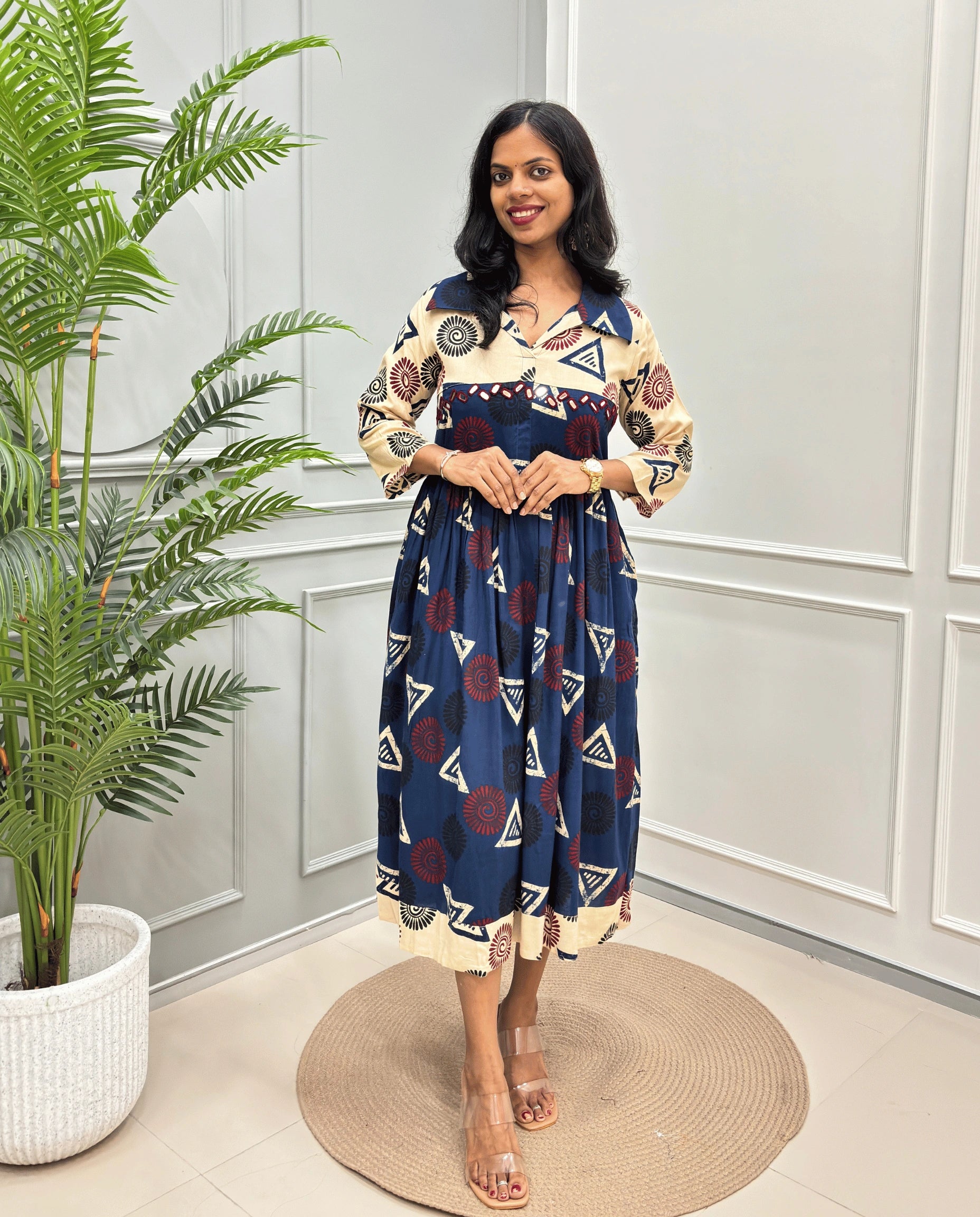 Navy-Blue Women's Ajrakh Vegetable dyed plant based Cotton Long-Kurti with a navy blue neck and cuffs, and a cream-colored dress featuring a geometric pattern of triangles and spiral in red, black, and blue, standing on a round woven rug in front of a white paneled wall with a green potted plant on the left.