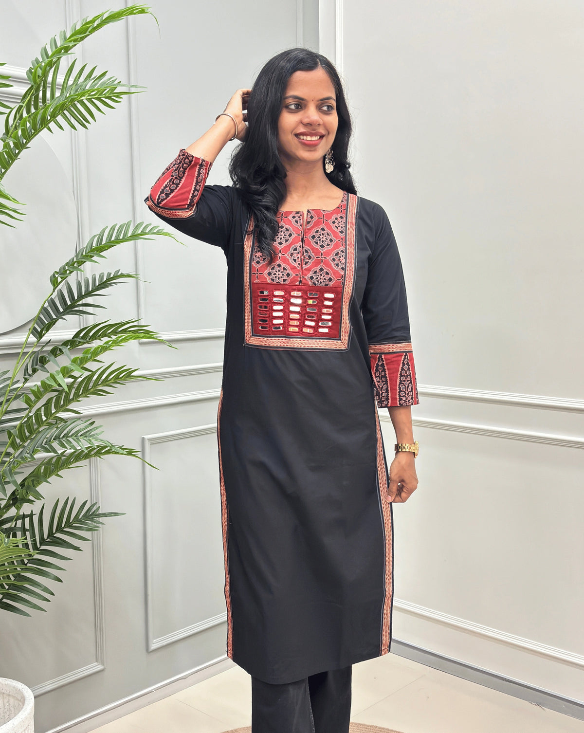 Front View of Black Women's Hand-block Ajrakh Pure Cotton Long Kurti with a red base and abstract Black & Beige geometric embroidery on the chest and sleeve cuffs, Hand-mirror work on Front, Ajrakh print border on Sides & Front  paired with black Flared pants, standing in a decorated room with a potted plant.