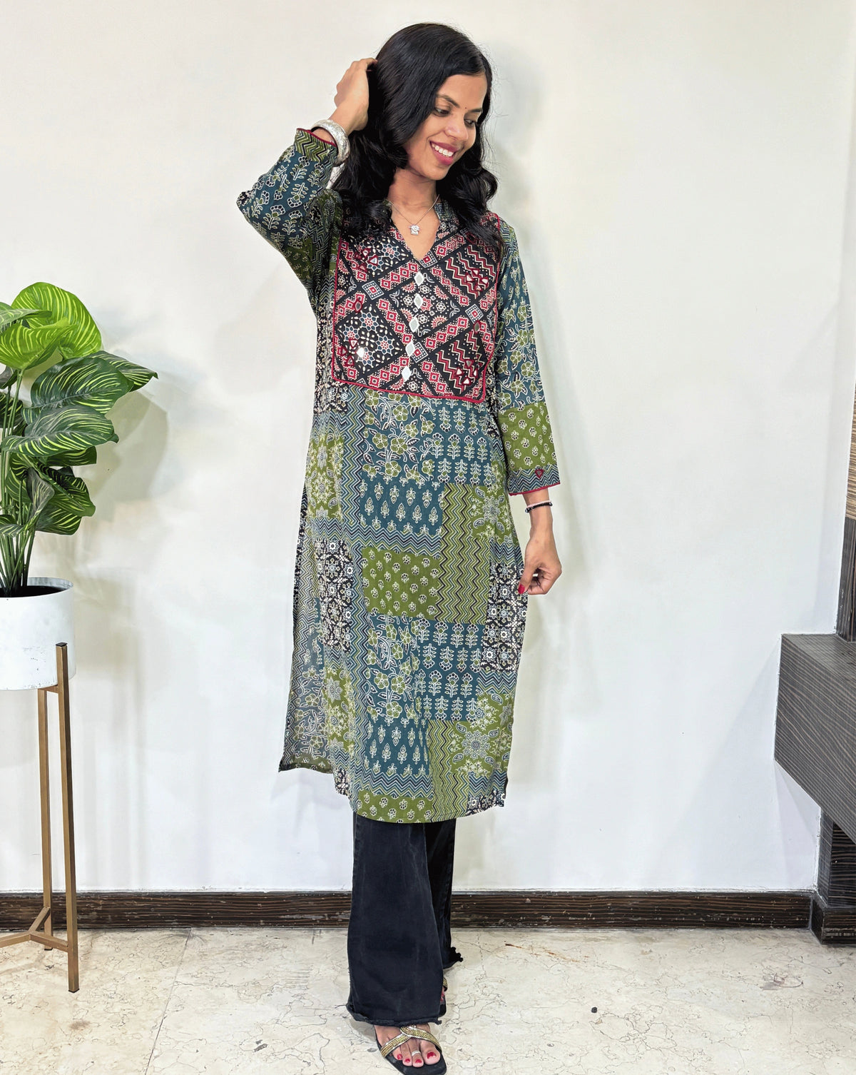 Green Women's Ajrakh Vegetable Dyed plant based cotton long kurti with green, black, and red patterns, paired with black flared pants, standing in a room with a white wall and a potted plant.