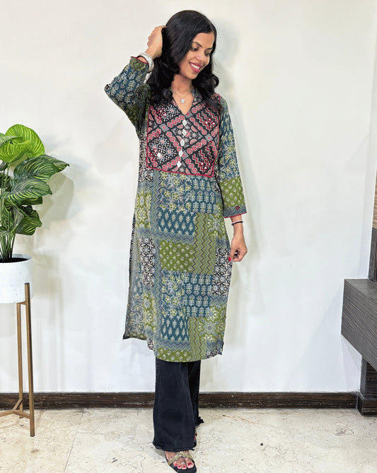 Green Women's Ajrakh Vegetable Dyed plant based cotton long kurti with green, black, and red patterns, paired with black flared pants, standing in a room with a white wall and a potted plant.