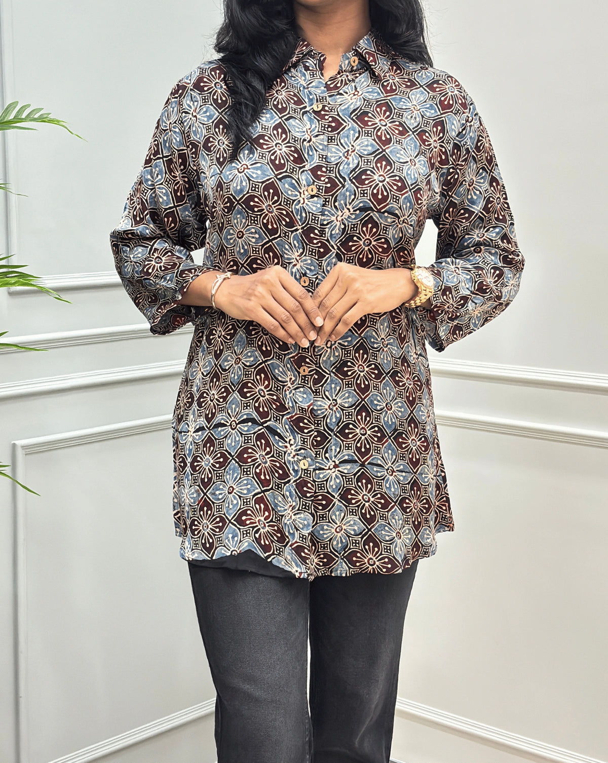 Close-up of Black Women's Ajrakh Pure Modal Silk Shirt with Full-Sleeves, with a Floral print on blue, and Red geometric pattern, black flared pants, standing on a round woven rug in front of a white paneled wall with a potted plant on the left