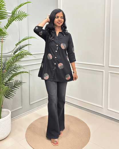Black Women's Ajrakh Vegetable dyed Plant based A-line cotton Short kurti with pink & white floral embroidery, paired with black flared jeans, standing on a round woven rug in a modern room with a potted plant.