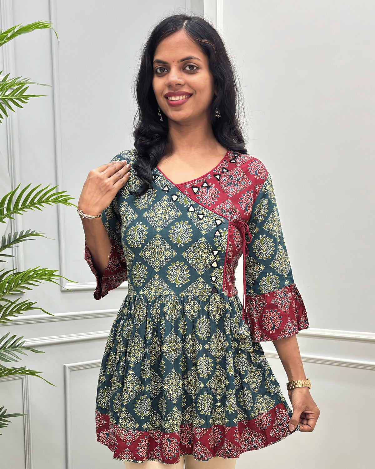 Close-up of Green women's Ajrakh Plant based cotton short-kurti with a Three-Quarter Sleeves (frill)  featuring Red & Green Geometric print ,  Angrakha Pattern with Hand-mirror work on V-shaped Neckline along with Lace, set against a light gray paneled wall.