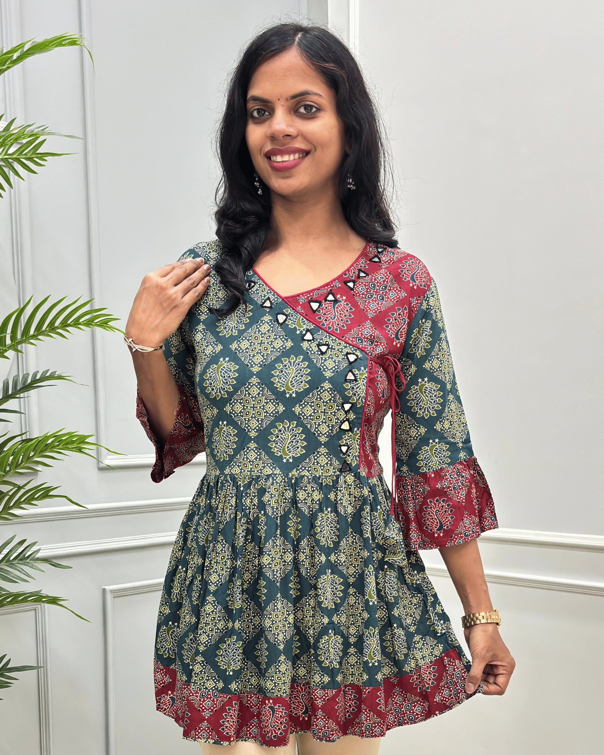 Close-up of Green women's Ajrakh Plant based cotton short-kurti with a Three-Quarter Sleeves (frill)  featuring Red & Green Geometric print ,  Angrakha Pattern with Hand-mirror work on V-shaped Neckline along with Lace, set against a light gray paneled wall.