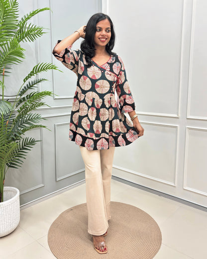 Green Women's Ajrakh Vegetable dyed plant based Cotton Short-Kurti with three-quarter frill sleeves, and waist to bottom,  paired with beige flared pant , standing on a round woven rug in front of a white paneled wall with a potted plant on the left.