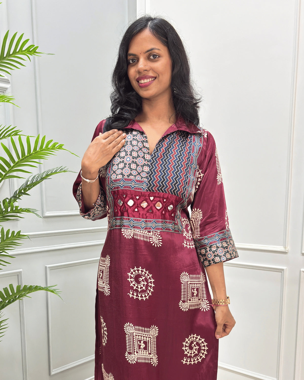 Close-up of Maroon Ajrakh Mashru silk Long kurti with multicolor geometric , warli print on front & sleeves along eith hand -mirror work on front , Collar V shaped neck patterns, standing in front of a white paneled wall with a green plant on the left.