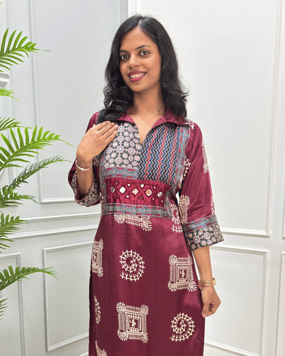 Close-up of Maroon Ajrakh Mashru silk Long kurti with multicolor geometric , warli print on front & sleeves along eith hand -mirror work on front , Collar V shaped neck patterns, standing in front of a white paneled wall with a green plant on the left.