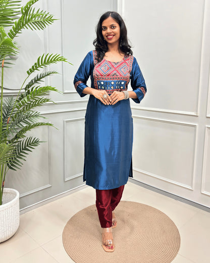 Blue Women's Ajrakh Mahru Silk Long-kurti with a geometric Print yoke in blue, red, and white, with Hand-Mirror Work,  paired with black pants, standing on a round woven mat in front of a white paneled wall with a potted plant.