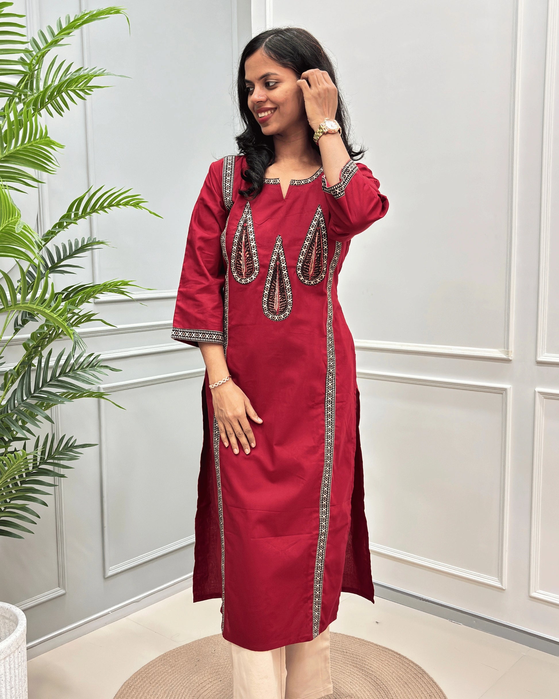 Front-view of Red Women's Hand-block Ajrakh Pure Cotton Long Kurti with Grey and Black embroidered details on the V-neckline and borders, standing in a decorated room with a potted plant.
