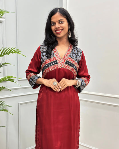 Close-up of Deep-Red Ajrakh Model silk fabric with three-quarter sleeves with intricate Red and Black patterned embroidery on the V-neckline, chest panel, and cuffs, standing in a light-colored room. 