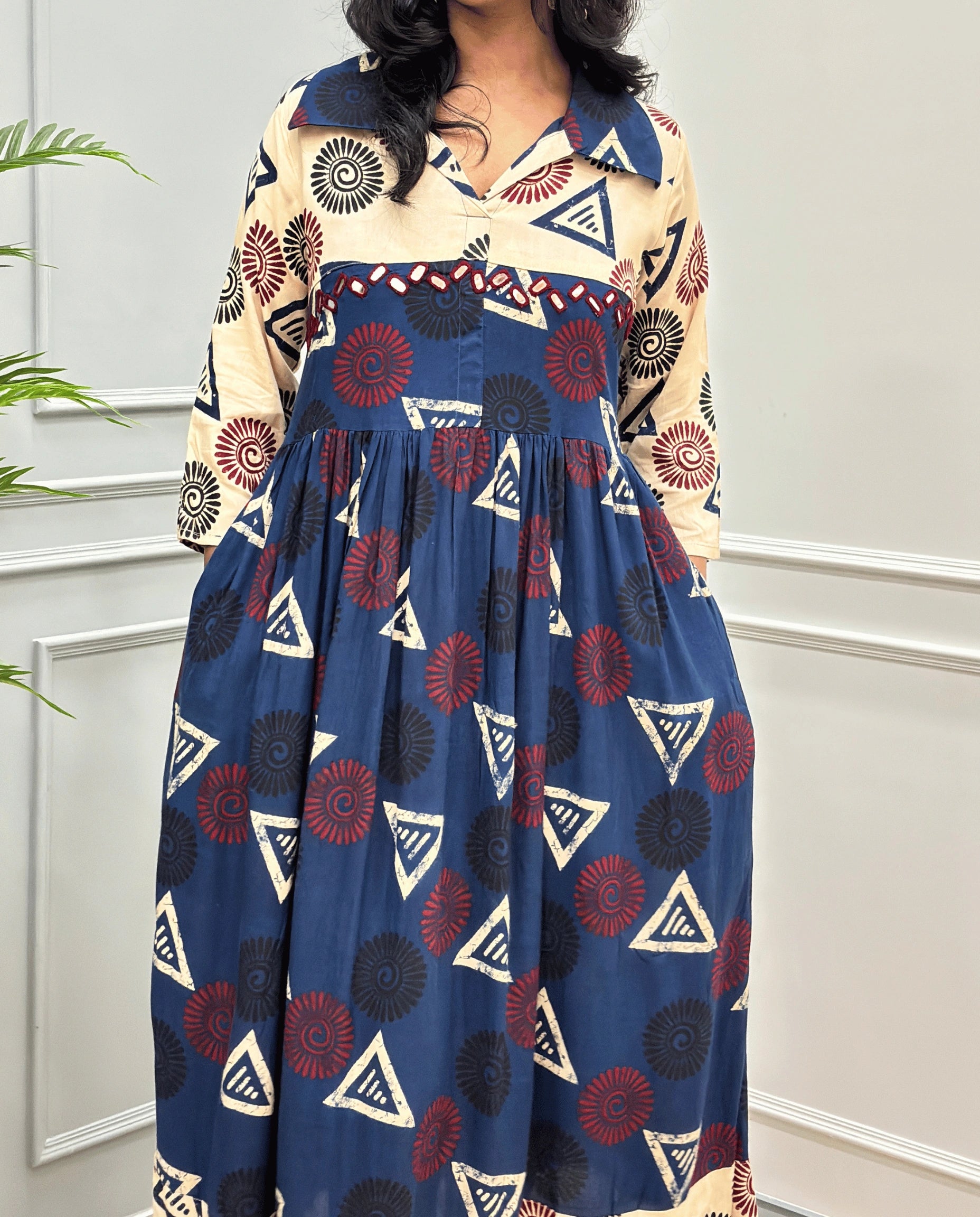 Navy-Blue Women's Ajrakh Vegetable dyed plant based Cotton Long-Kurti Featured with Three-Quarter Sleeves with a Beige neck and cuffs, and a Blue-colored collar & dress featuring a geometric pattern of triangles and spiral in Red, black, and beige, Hand-mirror work on front, standing on a round woven rug in front of a white paneled wall with a green potted plant on the left.