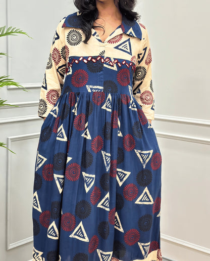 Navy-Blue Women's Ajrakh Vegetable dyed plant based Cotton Long-Kurti Featured with Three-Quarter Sleeves with a Beige neck and cuffs, and a Blue-colored collar & dress featuring a geometric pattern of triangles and spiral in Red, black, and beige, Hand-mirror work on front, standing on a round woven rug in front of a white paneled wall with a green potted plant on the left.
