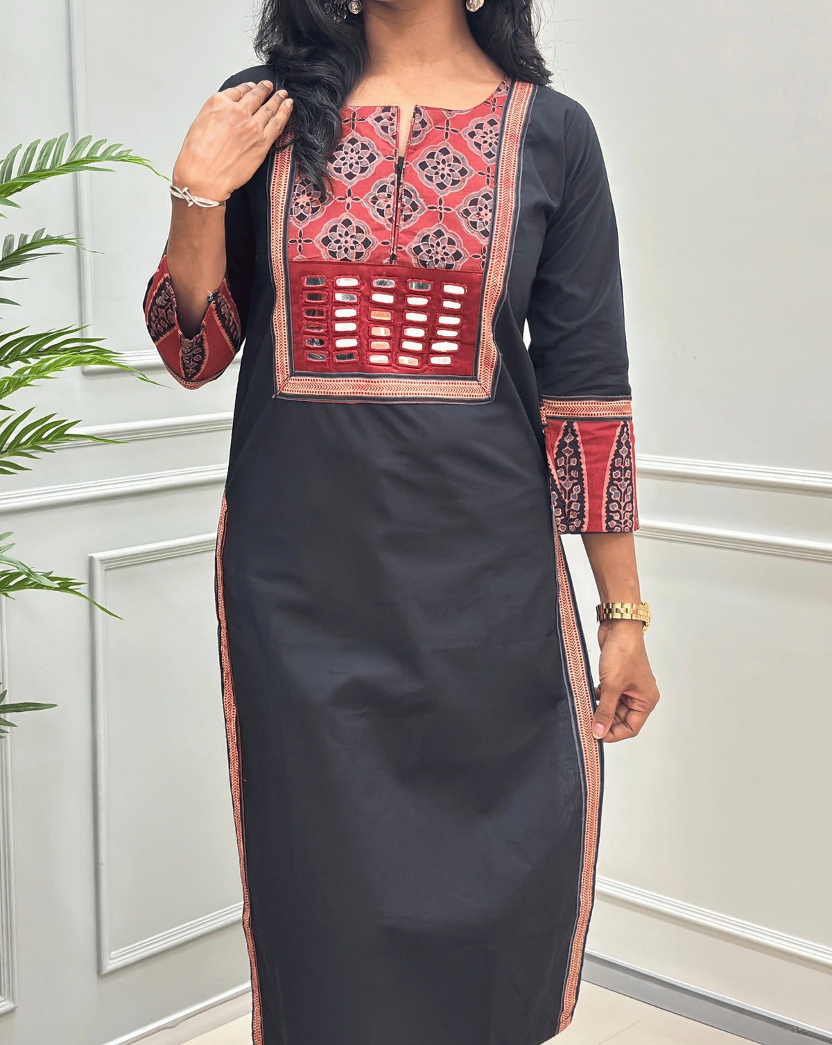 Close-up of Black Women's Hand-block Ajrakh Pure Cotton Long Kurti Featured with Three-quarter Sleeves & Round neck with V-shape cut with a red base and abstract Black & Beige geometric embroidery on the chest and sleeve cuffs, Hand-mirror work on Front, Ajrakh print border on Sides & Front, with Side Pockets., paired with black Flared pants, standing in a decorated room with a potted plant.