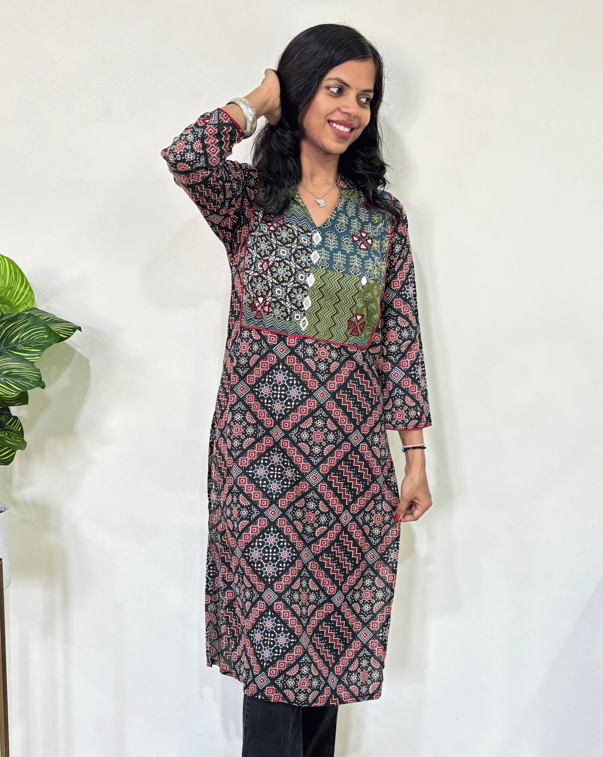 Front view of Black Women's Ajrakh Vegetable Dyed plant based cotton long kurti with green, black, and red patterns, Hand-mirror work on front & Sleeves paired with black flared pants, standing in a room with a white wall and a potted plant.
