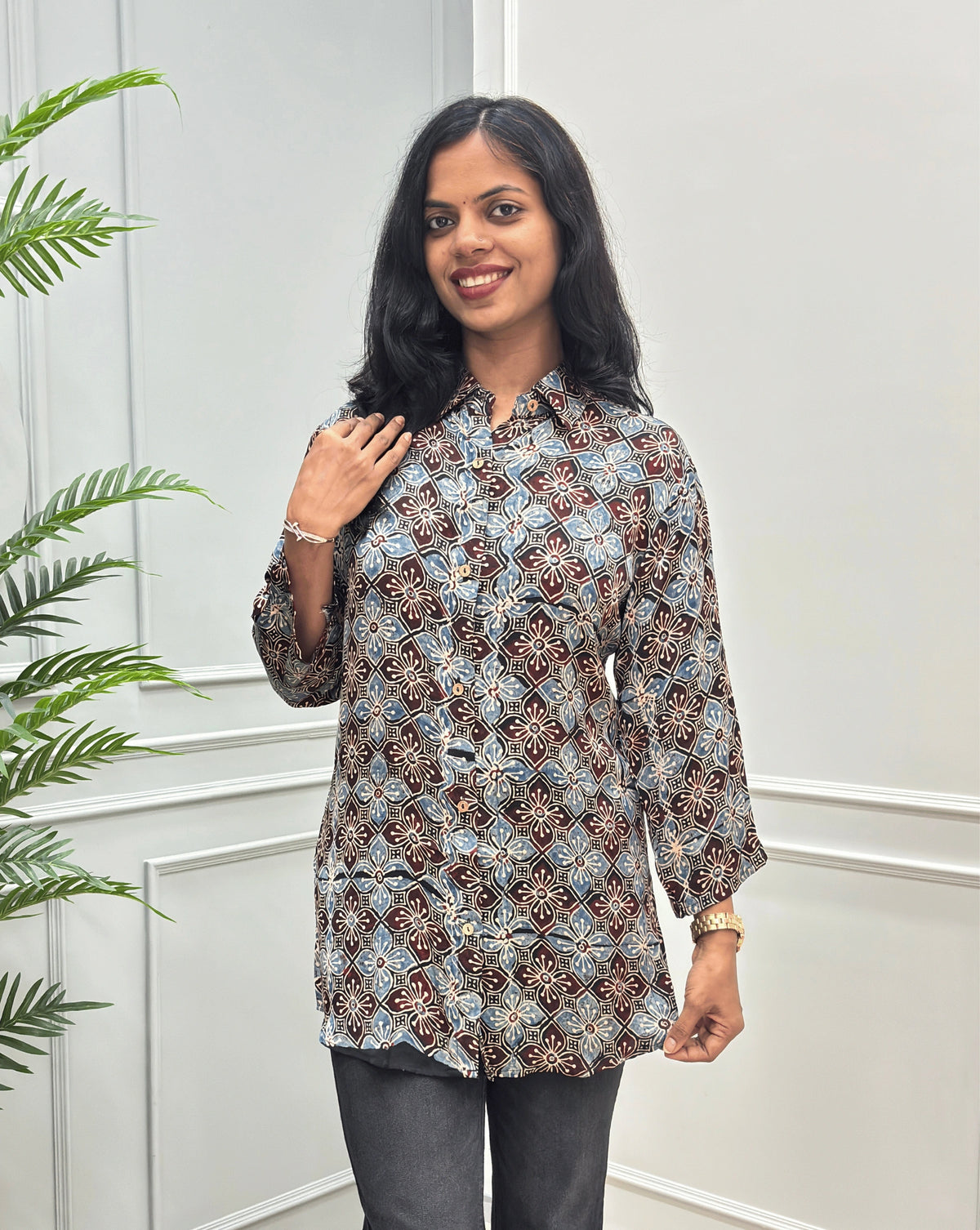 Front View of Black Women's Ajrakh Pure Modal Silk Shirt with a Floral print on blue, and Red geometric pattern, black flared pants, standing on a round woven rug in front of a white paneled wall with a potted plant on the left