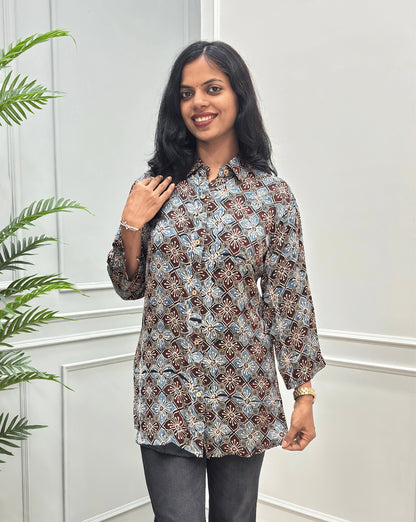 Front View of Black Women's Ajrakh Pure Modal Silk Shirt with a Floral print on blue, and Red geometric pattern, black flared pants, standing on a round woven rug in front of a white paneled wall with a potted plant on the left