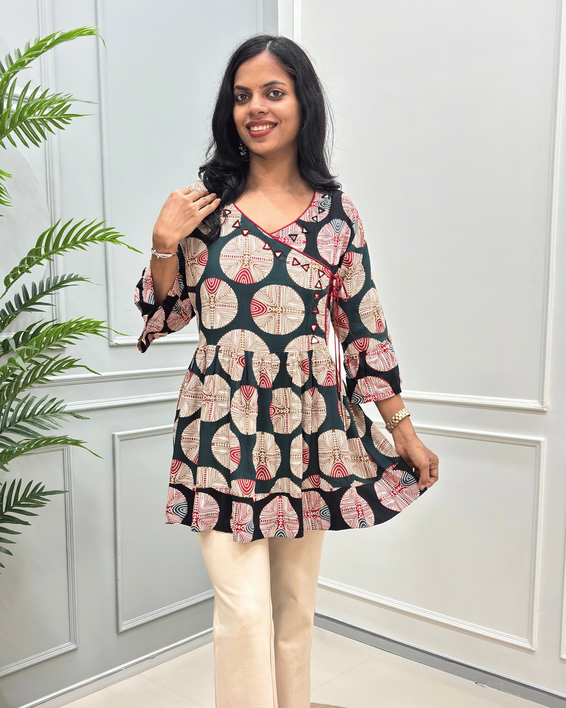 Front view of Green women's Ajrakh Plant based cotton Short-Kurti with a multicolor abstract print of circles and geometric patterns, Black color base on top bottom & sleeves cuffs in paired with beige pants, standing in front of a white paneled wall.