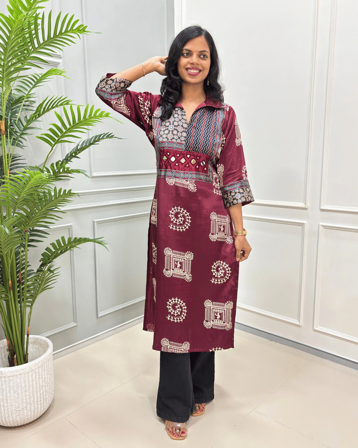 Maroon women's Pure Mashru Silk Long Kurti with red , Black & white geometric patterns on the yoke and sleeves, paired with black Flared pants, standing in a room with a potted plant.