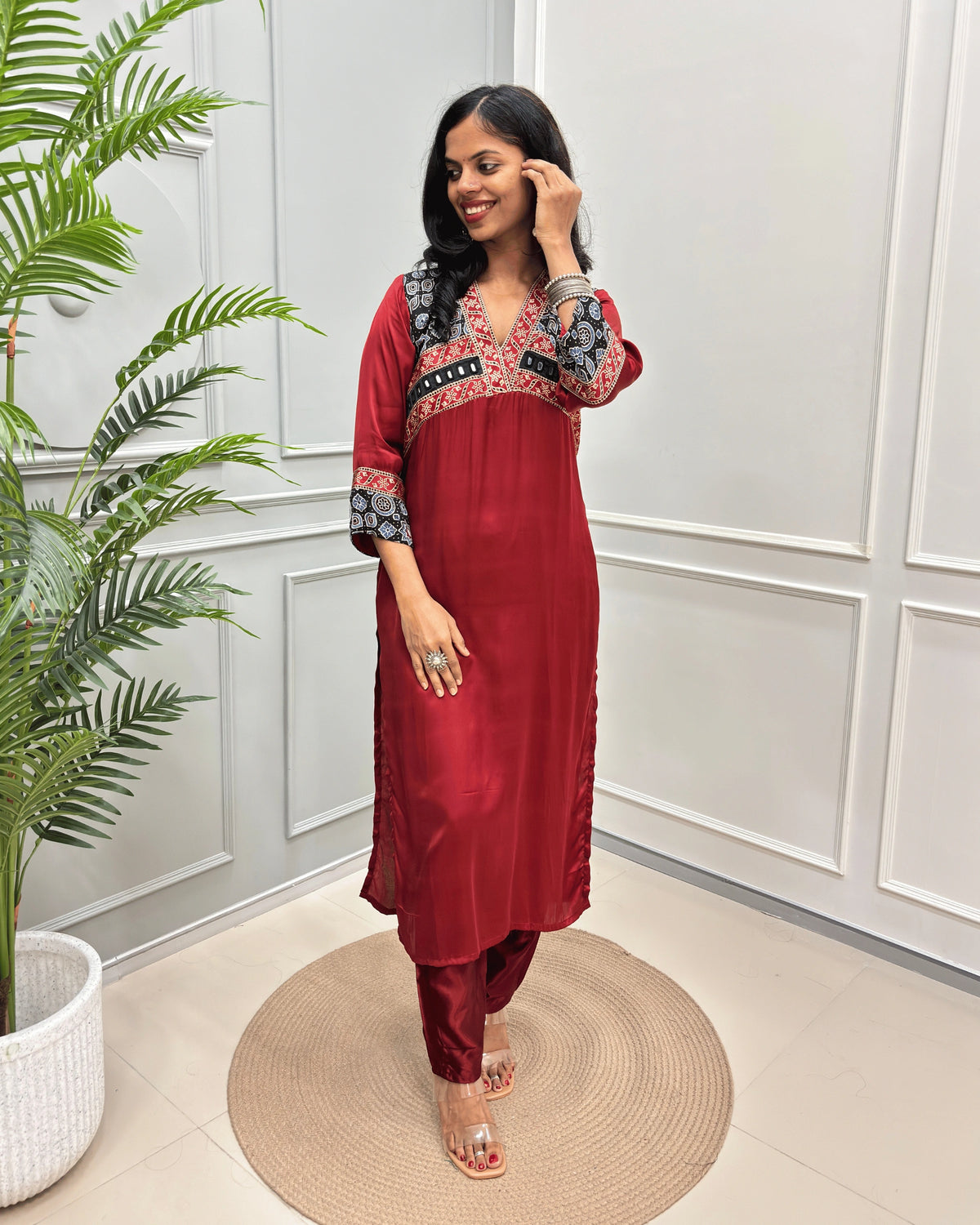 Deep Red Women's Ajrakh Modal Silk Long-Kurti with intricate bkack base & white & red Ajrakh print on the yoke and cuffs, paired with black pants and clear heeled sandals, standing on a round woven mat in a decorated room.
