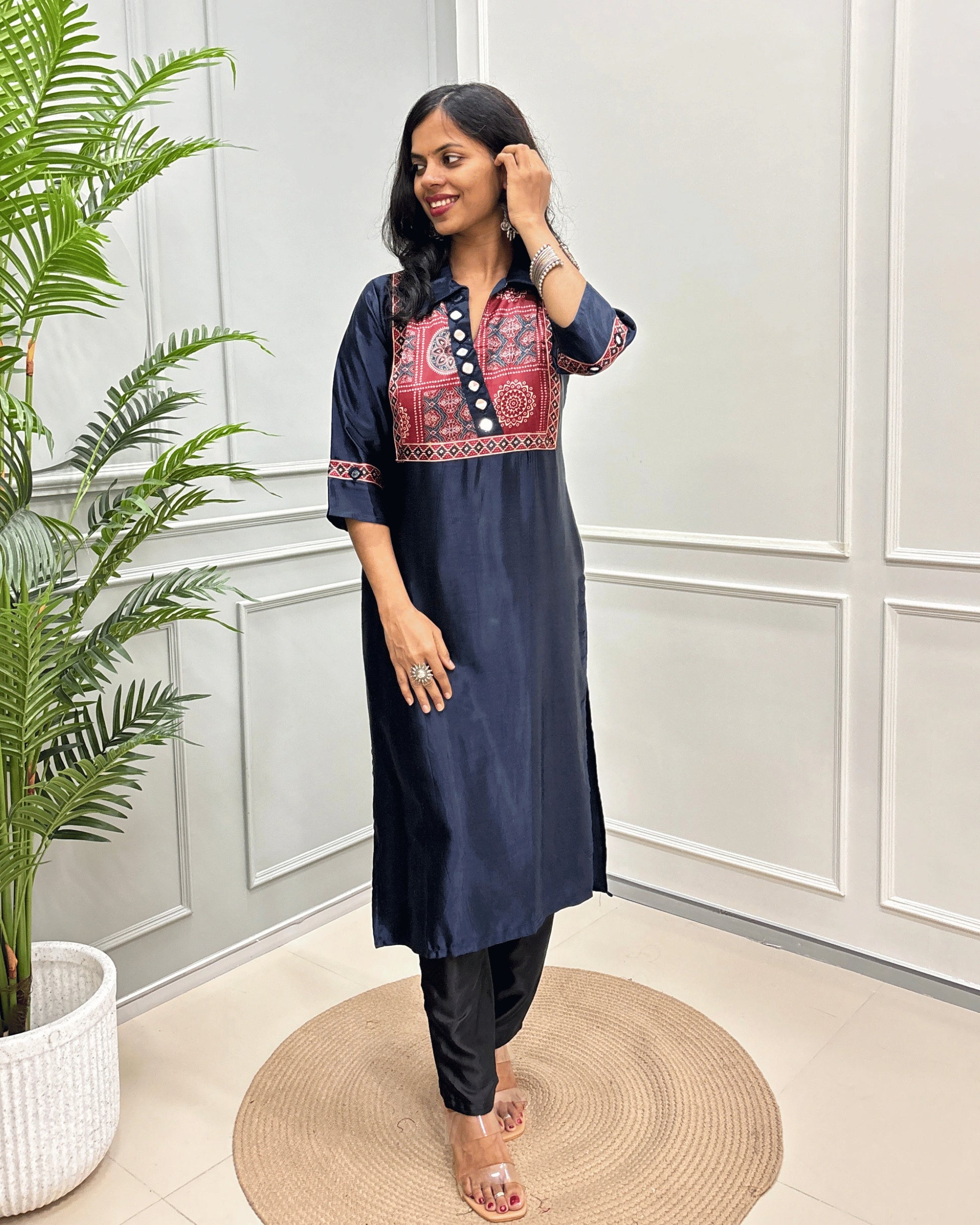 Navy-Blue Women's Ajrakh Mashru Silk Long Kurti with colorful embroidered detailing on the chest and sleeves, paired with black pants, standing in a decorated room with a potted plant and paneled wall.