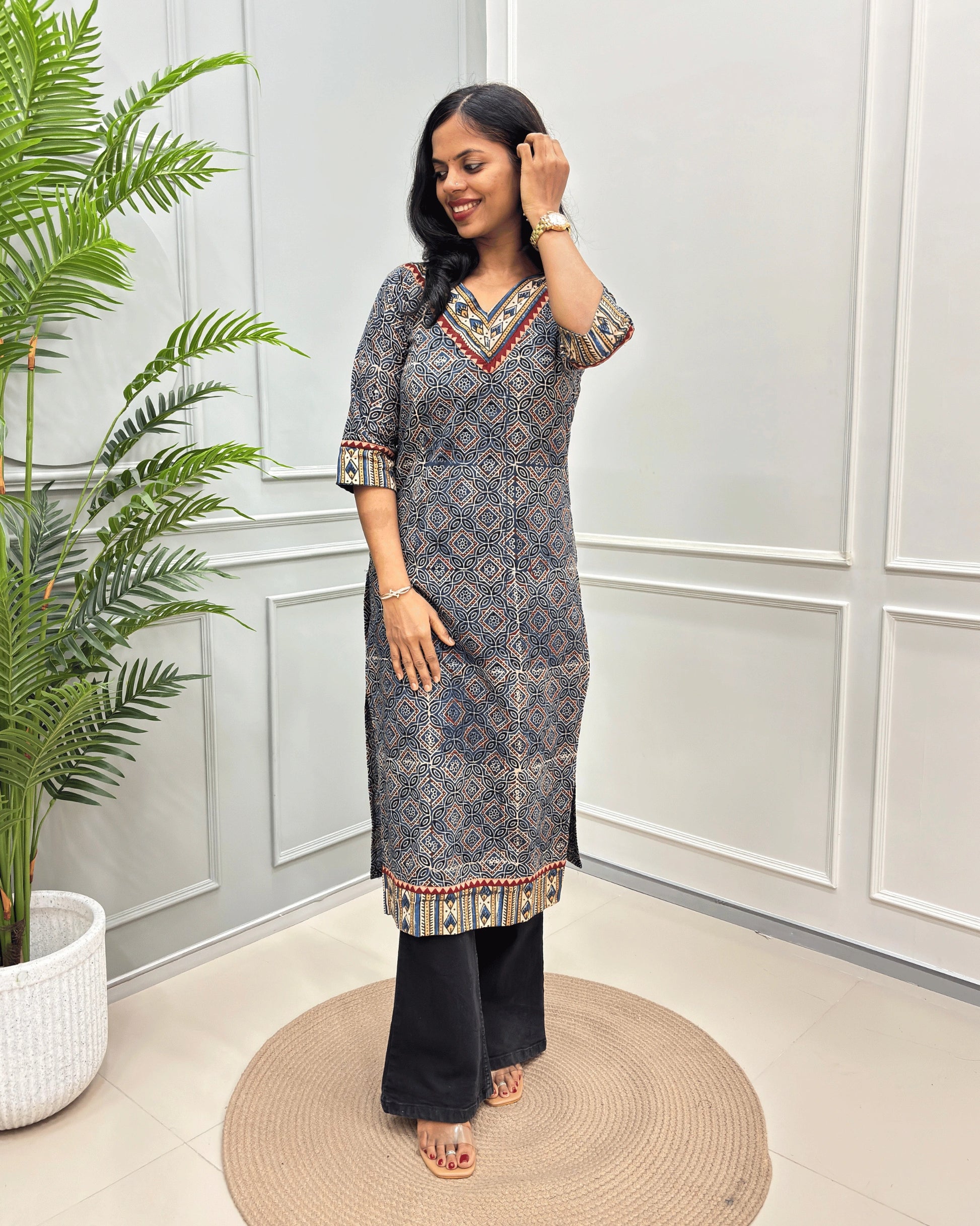 Blue Women's Ajrakh Hand-block Pure Cotton Long-Kurti with red and Beige geometric patterns, paired with Black Flared pants, standing on a round woven mat in front of a white paneled wall with a green potted plant.