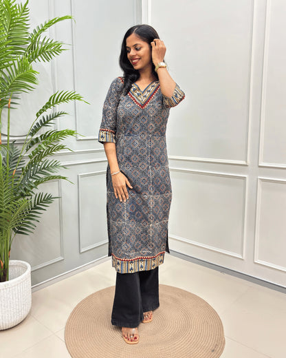 Blue Women's Ajrakh Hand-block Pure Cotton Long-Kurti with red and Beige geometric patterns, paired with Black Flared pants, standing on a round woven mat in front of a white paneled wall with a green potted plant.