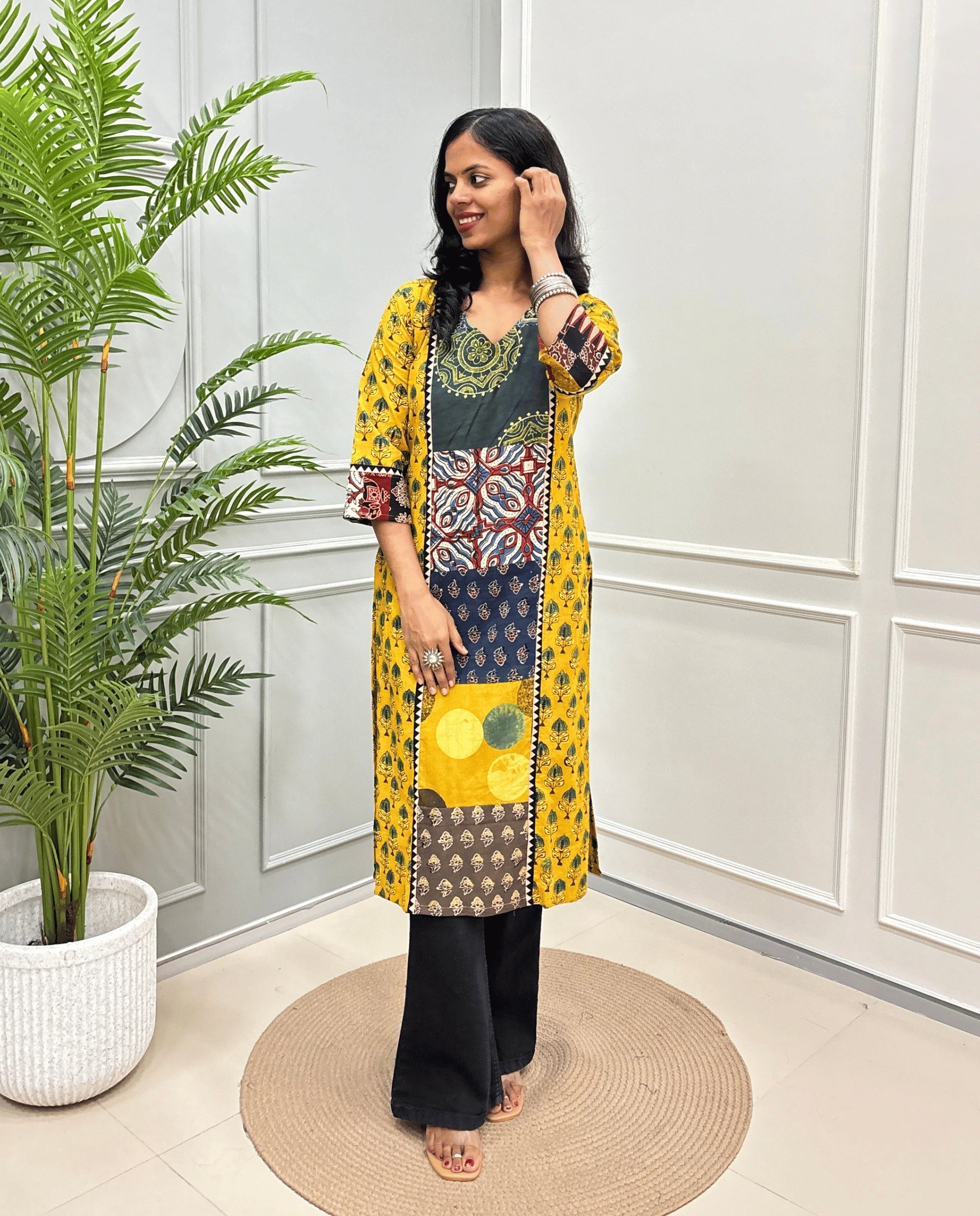 Yellow women's Hand-block Ajrakh  pure cotton Long Kurti with three-quarter sleeves, Ajrakh print on front and sleeves and black wide-leg pants, standing on a round woven mat in front of a white paneled wall next to a potted palm.