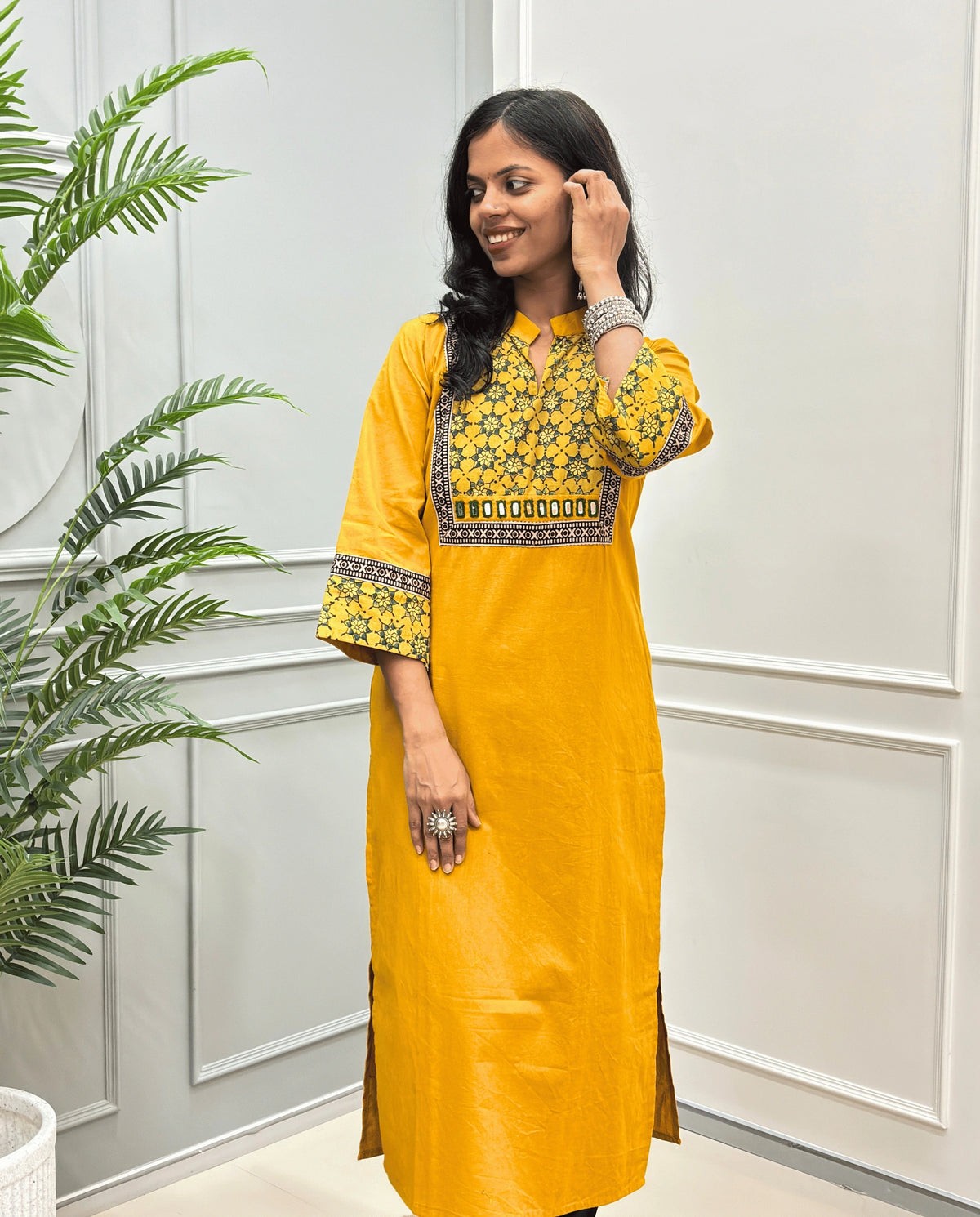 Front view of Yellow Women's Hand-block Ajrakh Pure Cotton Long kurti with intricate green geometric print on the chest and sleeve cuffs, hand-mirror work on front , paired with black pants , standing on a round woven mat in front of a white paneled wall with a potted plant.