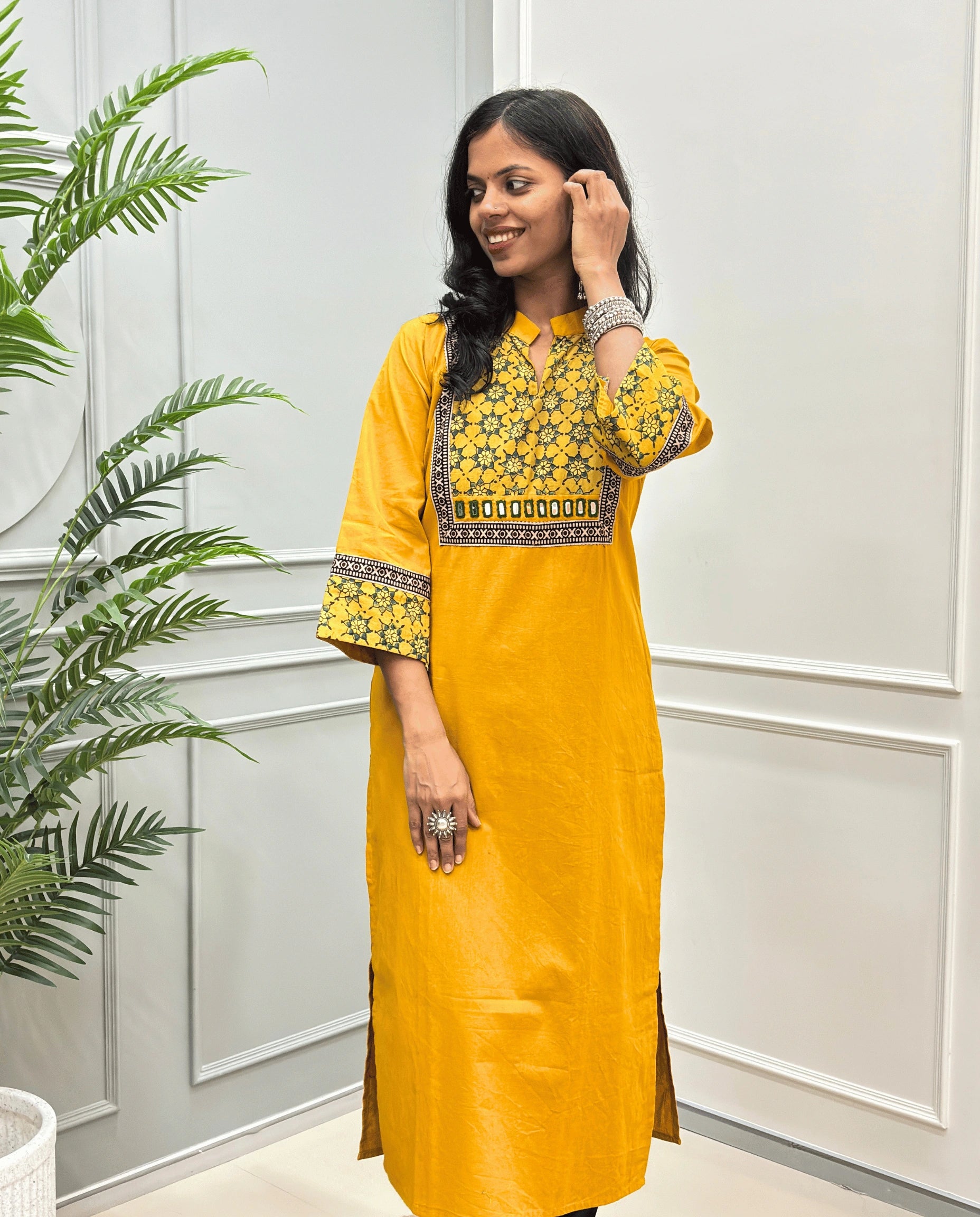 Front view of Yellow Women's Hand-block Ajrakh Pure Cotton Long kurti with intricate green geometric print on the chest and sleeve cuffs, hand-mirror work on front , paired with black pants , standing on a round woven mat in front of a white paneled wall with a potted plant.