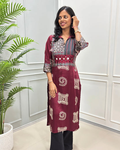 Front view of Maroon Ajrakh pure Mashru Silk Long kurti with geometric print in red, white, and beige on Front & sleeves, standing in a bright room with a white paneled wall and a potted plant.