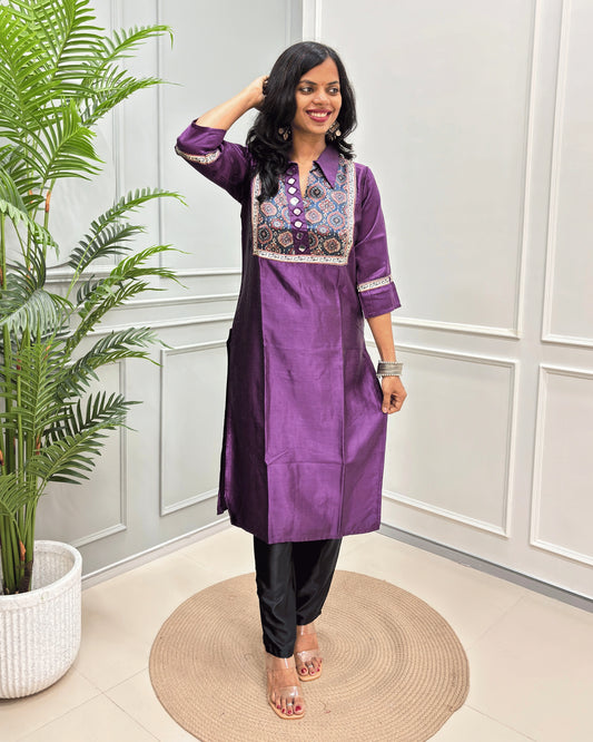 Purple Women's Ajrakh Mashru Silk Long Kurti with colorful embroidered detailing on the chest and sleeves, paired with black pants, standing in a decorated room with a potted plant and paneled wall.