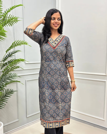 Front View of Blue Women's Ajrakh Hand-block Cotton Long Kurti with red, Blue and Beige ajrakh Print on V-neck ,Strip print on trim & Sleeves , standing in front of a white paneled wall with a green plant on the left.